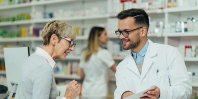 Young male pharmacist giving prescription medications to senior female customer in a pharmacy with female pharmacist in the background